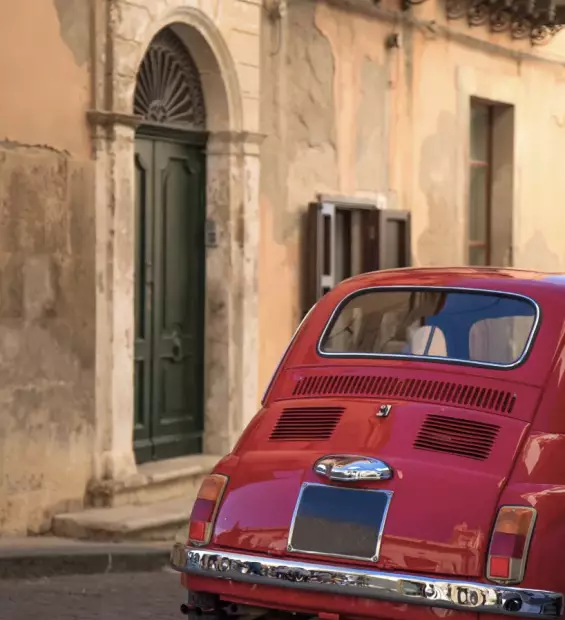 Rear view of a vintage red Fiat 500 parked on a stone street next to an old building with a classic green wooden door.